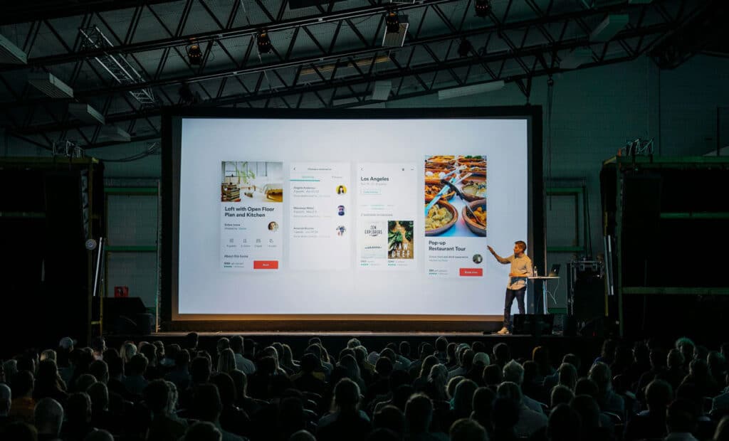 Speaker presenting a mobile app concept to an audience in a dimly lit auditorium.